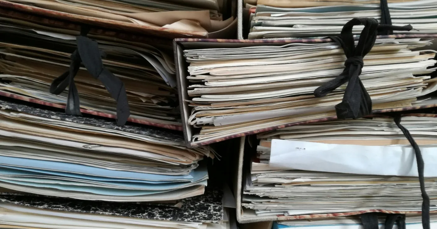 Piles de dossiers papier serrés par des rubans noirs, dans une réserve d’archives.